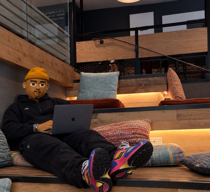 A person wearing a yellow beanie and black tracksuit (with face obscured by emoji) works on an Apple MacBook while reclining on wooden steps with colorful cushions in a modern interior space.
