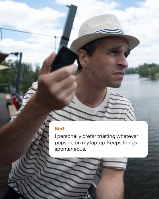A man in a striped shirt and straw hat holding up a black device outdoors near water, with an overlaid message from 'Bert' reading 'I personally prefer trusting whatever pops up on my laptop. Keeps things spontaneous.'