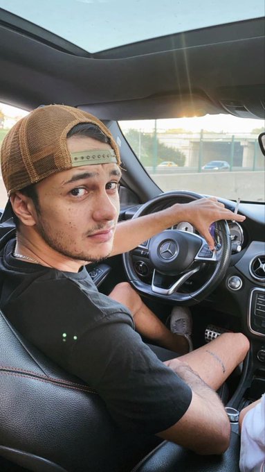 A young man wearing a tan backwards cap and black t-shirt sits in the driver's seat of a Mercedes-Benz, looking back over his shoulder toward the camera.