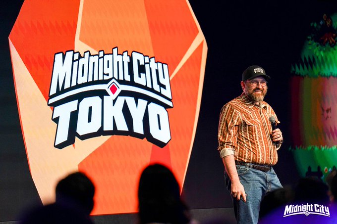 A bearded man in an orange striped shirt and a cap speaks on stage holding a microphone, with a large 'Midnight City TOKYO' logo displayed on the screen behind him.