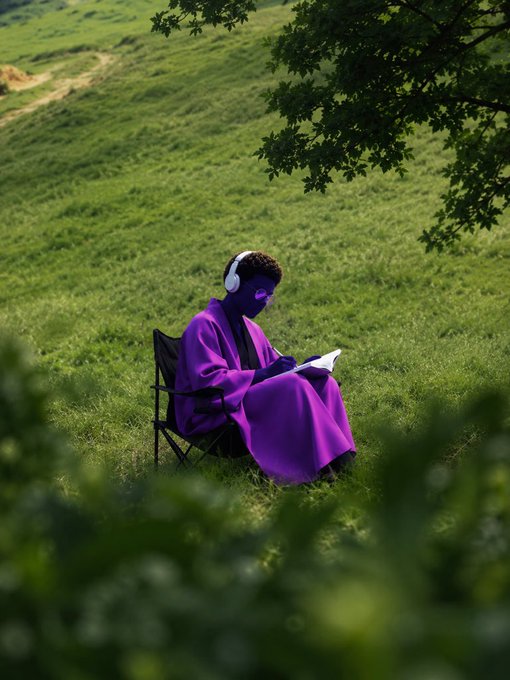 A person with purple-tinted skin wearing a purple blanket and white headphones, sitting on a folding chair in a green grassy field, sketching in a notebook under a tree.