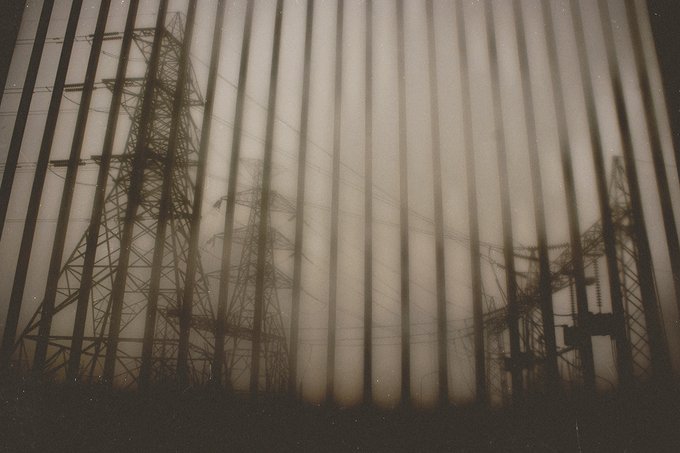 Silhouettes of electrical transmission towers and power lines viewed through vertical blinds or slats, creating a layered, hazy effect.