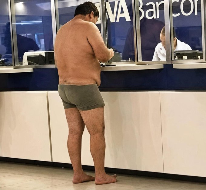 A shirtless man in gray underwear stands barefoot at a "NVA Bancontact" bank counter, interacting with a teller through the glass window.