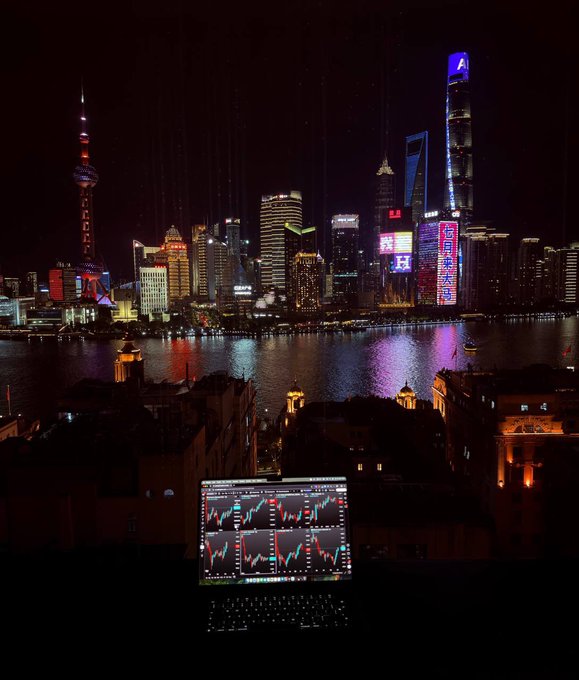 A nighttime photograph of the Shanghai skyline featuring illuminated skyscrapers and neon signs across the Huangpu River, with a laptop displaying financial trading charts in the foreground.