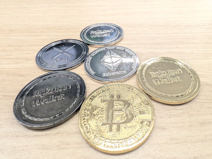 A collection of five metallic cryptocurrency commemorative coins rests on a wooden surface, featuring replicas of Bitcoin (BTC), Ethereum (ETH), and XRP, along with three coins branded with the "Rakuten Wallet" logo, representing the exchange's "Real Coin" series before the addition of their upcoming Shiba Inu (SHIB) coin.