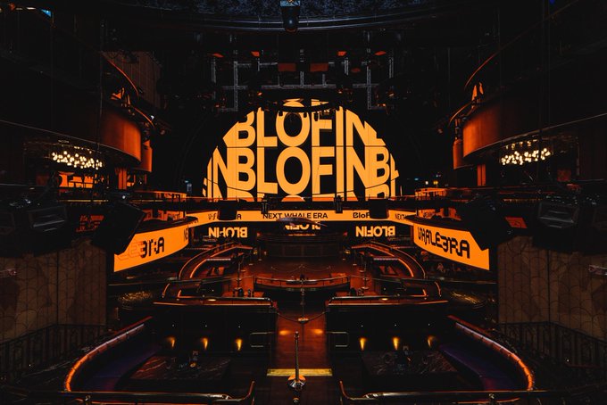 A dimly lit nightclub interior bathed in orange lighting features large illuminated displays reading "BLOFIN BLOFIN" and "NEXT WHALE ERA" wrapping around the venue, with mirrored text effects visible on the surrounding structures. The image showcases the atmospheric setup of WHALE3RA at MARQUEE Singapore, aligning with the post's announcement of "The Next Whale Era" beginning tonight.