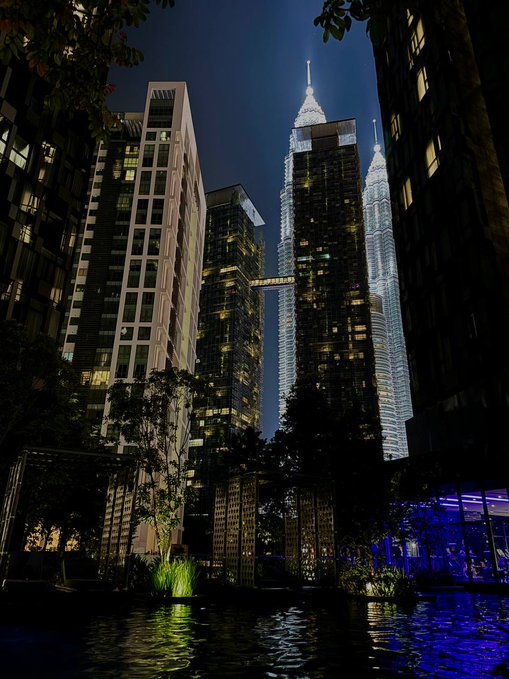 A nighttime view of illuminated skyscrapers, including the Petronas Twin Towers, rising above a dark water feature and lush plants.