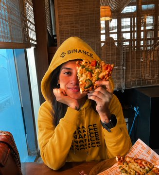 The image features a woman sitting at a table with two slices of pizza in front of her. She is wearing a yellow hoodie that says "Binance" on it, indicating that she might be a Bitcoin enthusiast or working for a company related to cryptocurrency. The table also has a cup and a bottle placed next to the pizzas.