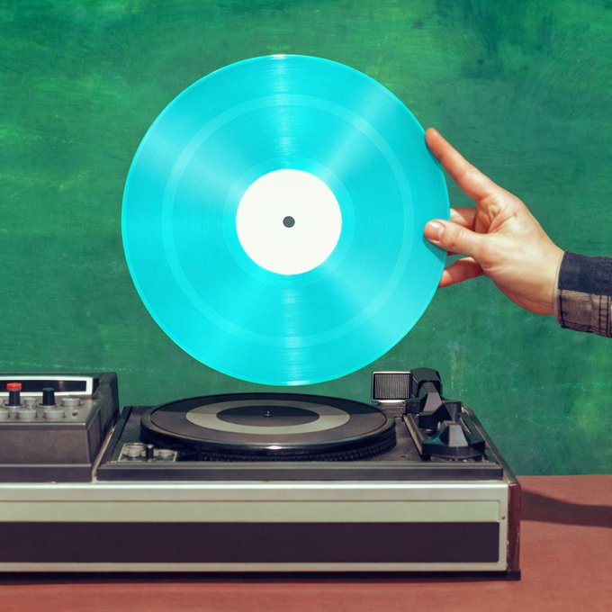 A hand holds up a bright turquoise vinyl record in front of a retro record player against a textured green background.
