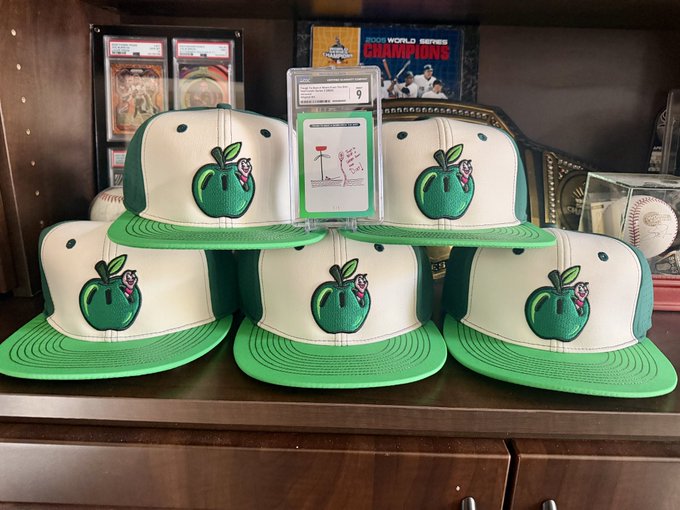 Five white and green baseball caps featuring an embroidered green apple logo are displayed on a wooden surface, with a graded sports card visible behind them.