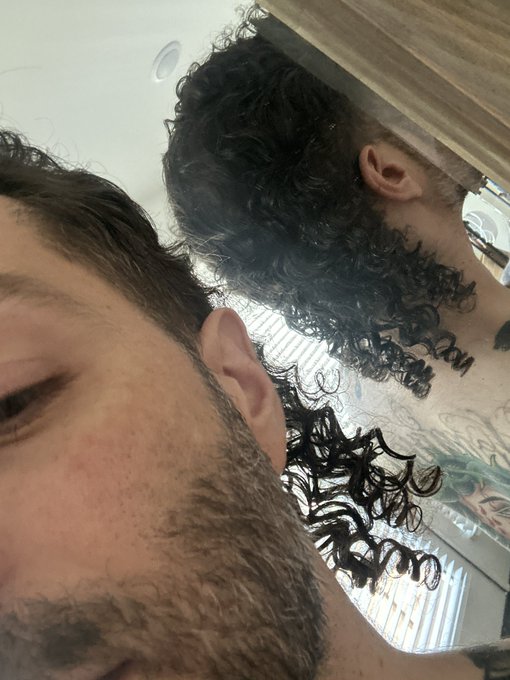 A man with curly hair and a full beard appears to be showcasing the back of another person's head, which features a voluminous curly mullet hairstyle. The image illustrates the "mullet maxxing" trend referenced in the caption, with the length and curls on the back perfectly embodying the bullish crypto-inspired commitment to rocking a mullet throughout the market run.
