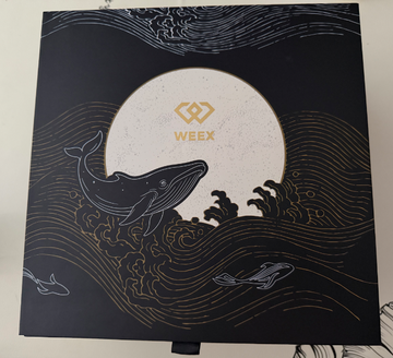 A dark blue box or notebook featuring gold artwork of whales swimming in waves under a moon, with the logo 'WEEX' in the center.