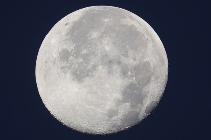 A close-up photograph of a full, bright gray moon against a dark blue sky, showing visible craters and lunar maria.