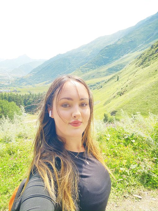 A selfie of a young woman with long brown hair wearing a black shirt, standing in a lush green mountain landscape.