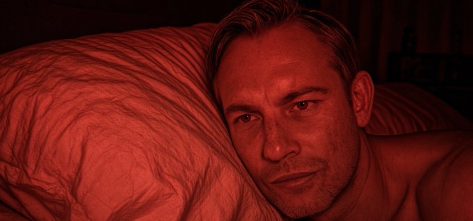 A man's face is illuminated by deep red lighting as he lies on a wrinkled pillow, gazing directly at the camera.
