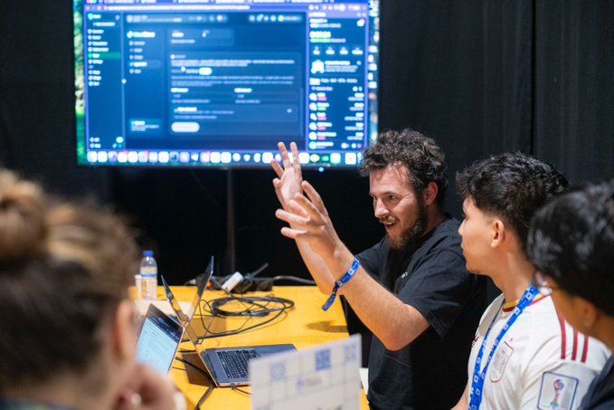 A man wearing a black shirt and a blue wristband gesturing with his hands while collaborating with two young men around a laptop in front of a large monitor displaying a code interface.