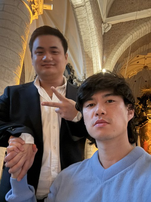 Two men pose for a selfie inside an ornate cathedral, with one wearing a black blazer over a white shirt and making a hand gesture, while the other wears a light blue sweater and looks seriously into the camera. The image appears to accompany a post about onboarding a newcomer, "Jeremy," into trading—visually illustrating the mentorship dynamic described in the caption.