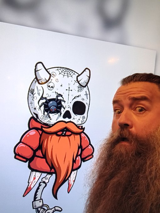 A man with a large brown beard poses next to a colorful illustration of a horned skull character with an orange beard, red hoodie, and a small bat emerging from its eye.
