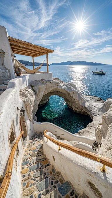 A scenic Mediterranean-style cove with whitewashed architecture, a stone staircase with wooden railings, a small cave opening to turquoise water, and a boat visible in the sunlit sea.