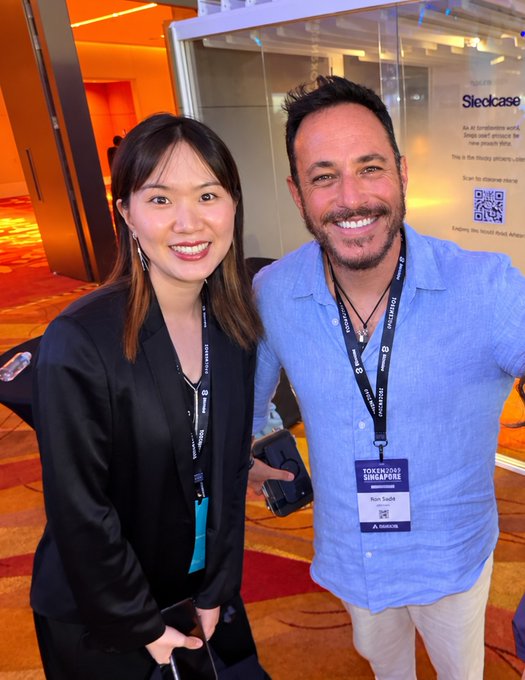 Two attendees at Token2049 Singapore pose for a photo; the man wears a blue shirt with a 'Ron Sade' name badge, and the woman wears a black blazer.