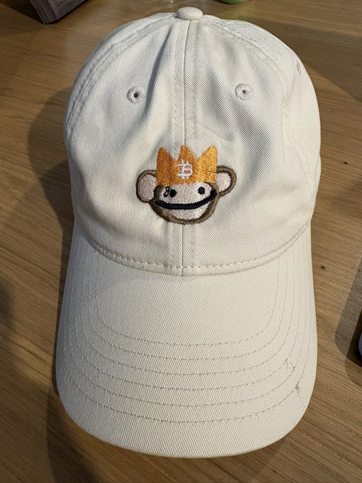 A cream-colored baseball cap rests on a wooden surface, featuring a small embroidered patch of a smiling monkey wearing an orange crown with the letter 'B'.