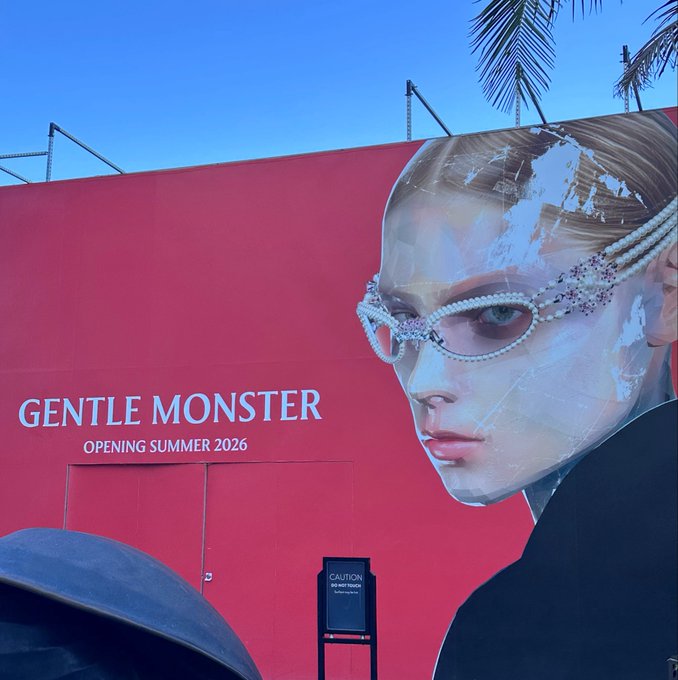 A red construction barrier with a 'GENTLE MONSTER OPENING SUMMER 2026' advertisement featuring a model wearing jeweled sunglasses, set against palm trees and a clear sky.