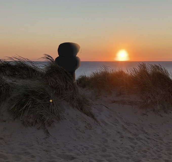 A large dark stone sculpture sits on sandy dunes surrounded by swaying beach grass, silhouetted against a glowing sunset over the calm sea, capturing a peaceful end to the day. The serene twilight scene perfectly matches the warm, relaxed "good evening frens" greeting.