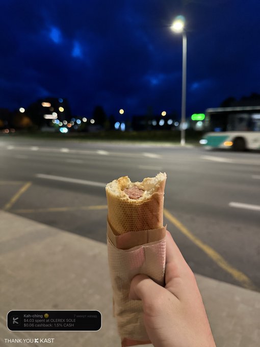 The image shows a person holding a hot dog wrapped in paper while standing next to a street light at night. The photo has been tagged with the caption "Kah-ching @KASTxyz" which suggests that the person who took the picture is from Japan and wants to share their experience of enjoying a hot dog on the go during nighttime hours.