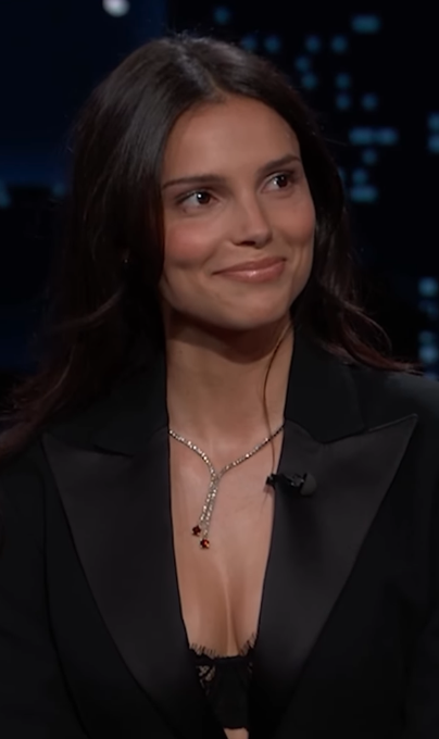 Emily Ratajkowski smiles during a talk show interview, wearing a black blazer and an elegant diamond necklace with a teardrop pendant. The image appears unrelated to the post, which discusses Inde Navarette and Anne Hathaway's Oscar odds on Polymarket.