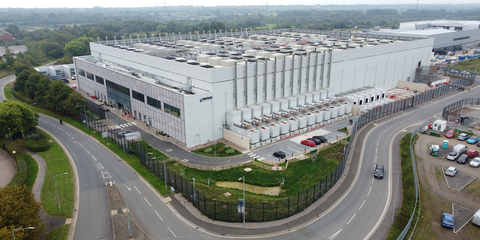 An aerial view of a large, modern data center complex with extensive rooftop cooling infrastructure, surrounded by roads and parking lots, illustrating the type of high-density AI facility where Nebius ($NBIS) has announced its deployment of NVIDIA-powered AI infrastructure at Vantage Data Centers' CWL1 campus in Newport, Wales.