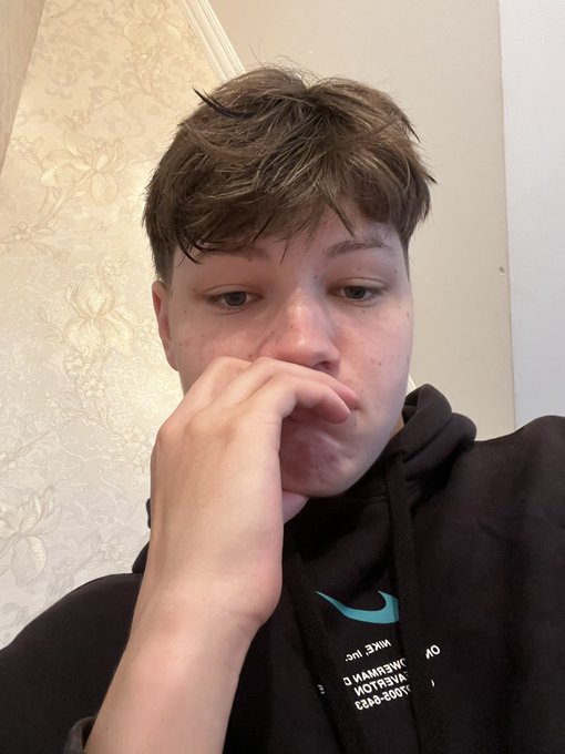A young man with brown hair takes a close-up selfie, resting his hand against his mouth in a contemplative pose while wearing a black Nike hoodie with reversed mirrored text visible on the chest.