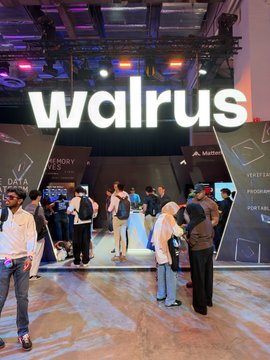 A photo of a convention or expo booth with a large illuminated "walrus" sign, showing people walking around an exhibition floor with "VERIFY" and "PROGRAM" signage visible.