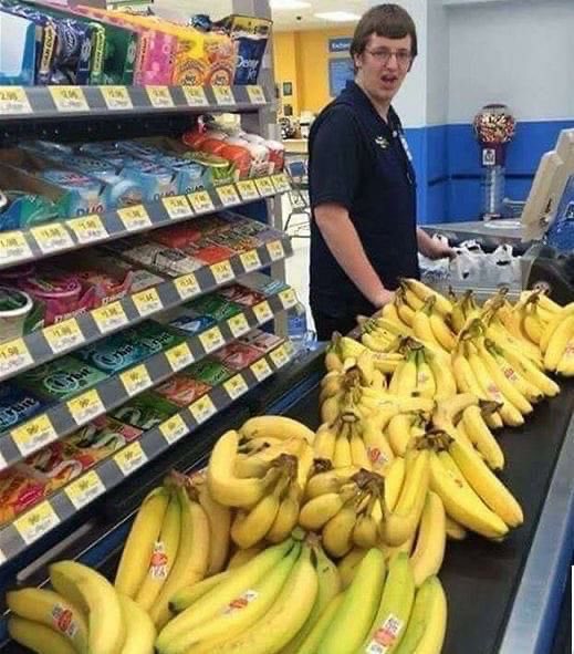 A store employee stands behind a display of fresh bananas at a grocery store checkout, with candy shelves visible on the left.