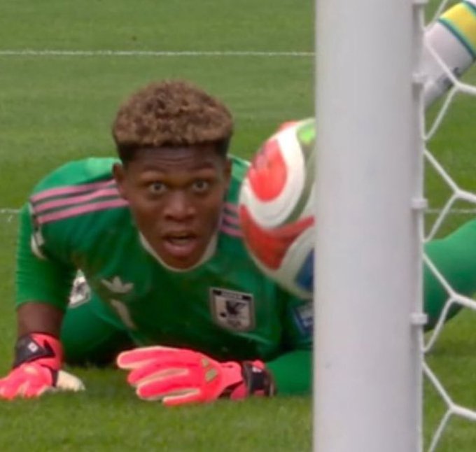 A goalkeeper wearing a green Mexico national team jersey with the number 11 and pink gloves dives toward a soccer ball near the goalpost.