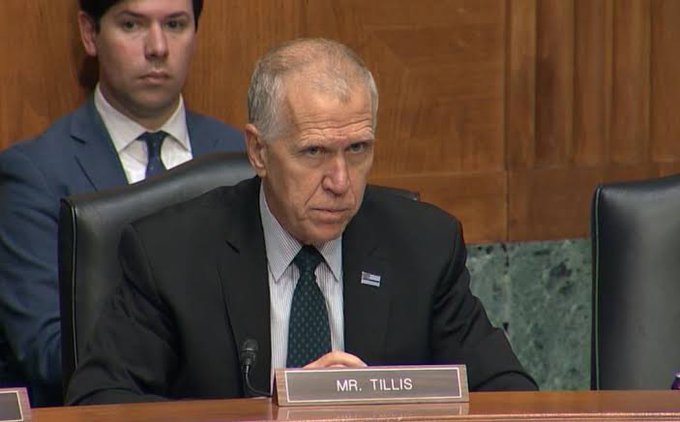 Senator Thom Tillis, identified by a "MR. TILLIS" nameplate, sits at a committee hearing with a serious expression, illustrating his remarks about the CLARITY Act's diminished chances amid midterm election pressures. A staff member stands behind him in the formal hearing room.