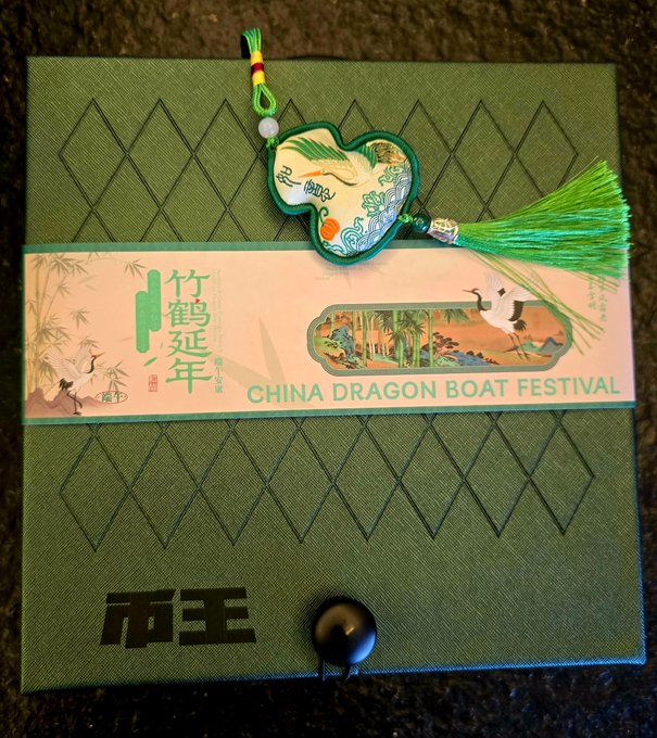 The image features a green box with an Asian writing on it, which appears to be related to a Chinese Dragon Boat Festival. The box contains a charm that has a dragon design on it, along with a tassel made of green thread hanging from it. There is also a small black button attached to the charm.