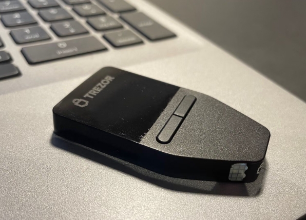 A Trezor hardware wallet rests on a laptop keyboard, representing device-level security for private keys—though the caption warns that user data breaches (names, emails, addresses) can still expose owners to targeted phishing and social engineering attacks despite the wallet's protection.