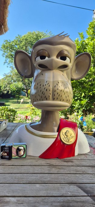 A painted sculpture of an ape character wearing a red sash with a gold medallion sits on a wooden table outdoors.