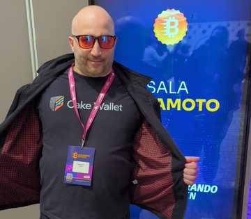 A man wearing red sunglasses and a black 'Cake Wallet' t-shirt opens his jacket to reveal the logo, standing in front of a Bitcoin promotional banner at what appears to be a crypto event.