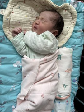 A photograph of a sleeping newborn baby swaddled in a light-colored floral blanket, resting on a blue patterned pillow or mat.