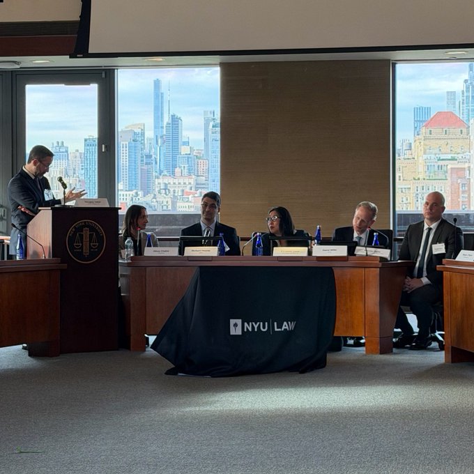 The image shows a group of people sitting around a table, engaged in a discussion or panel meeting. There are five individuals visible in the scene, with one person standing at a podium and another seated at the head of the table. The room has large windows that provide an expansive view of the city skyline outside.

Several bottles can be seen on the table, suggesting refreshments have been served during the event. A book is also present on the table, possibly containing relevant information or materials for the discussion.