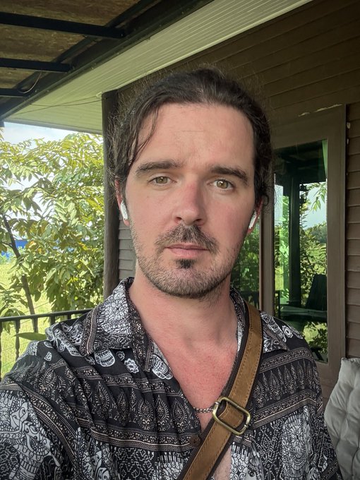 A selfie of a man with brown hair and a goatee wearing AirPods and a black-and-white patterned hoodie, standing on an outdoor balcony.