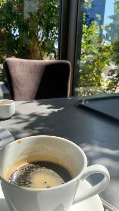 An empty white coffee cup with coffee residue sits on a table in a sunlit cafe, with a chair and a window view of green foliage in the background.