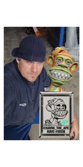 A man wearing a blue baseball cap and shirt poses with a green cartoon monkey character and a framed picture labeled 'HAVOC THE APE MAYC #18938'.