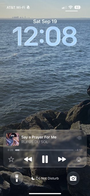 An iPhone lock screen shows the time 12:08 on Saturday, September 19, featuring a rocky coastline background and a music widget playing 'Say a Prayer For Me' by RUFUS DU SOL.