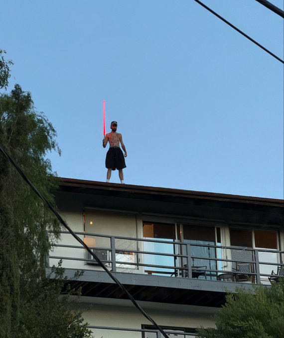 A shirtless man wearing a black kilt stands on the roof of a modern house at dusk, holding a glowing red lightsaber against a clear blue sky.