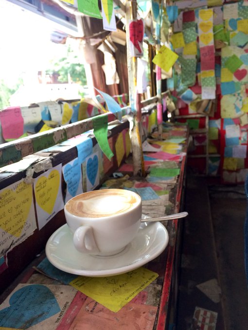 A cappuccino with latte art sits on a saucer amid a colorful display of handwritten sticky notes covering the walls and surfaces of a café, creating a cheerful, personal atmosphere that matches the upbeat "Good morning, hello Sunday" greeting.