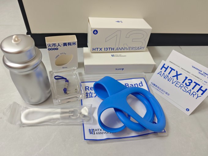 A collection of HTX 13th Anniversary promotional merchandise is arranged on a floor, including a silver bottle, a Resistance Band, and branded boxes.
