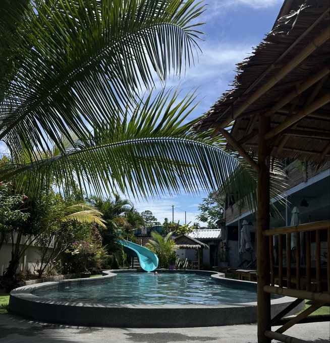 A tropical poolside escape featuring a turquoise pool with a winding green water slide, framed by swaying palm trees and a bamboo hut with a thatched roof—a serene offline retreat that captures the essence of luxury in disconnecting from the digital world.