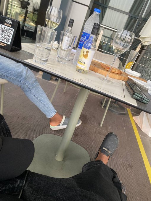 A first-person view of an outdoor café table with drinks including wine and water bottles, showing another person's legs in jeans and the photographer's feet in slippers, with a 'SCAN ME' QR code display on the table.